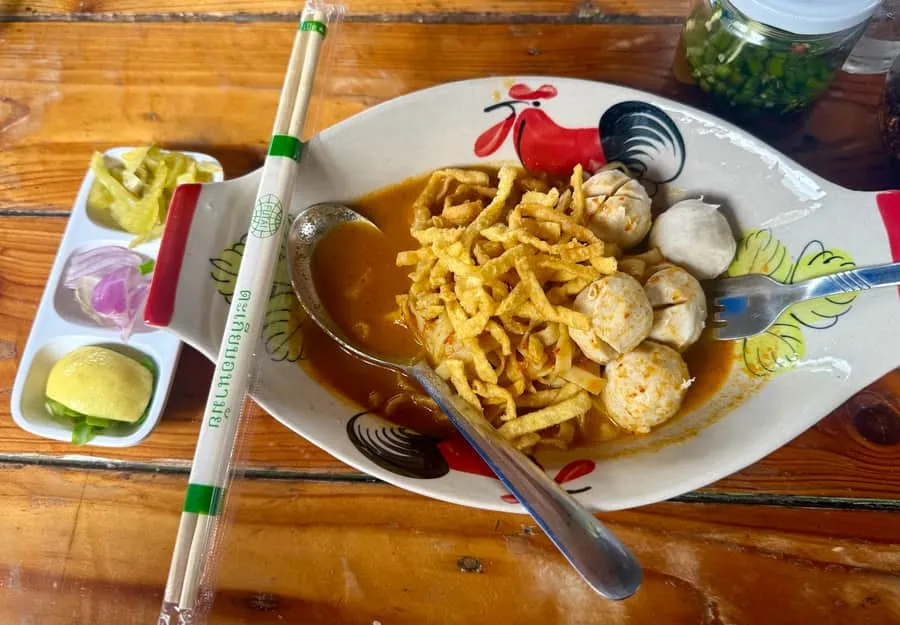 A bowl of khao soi with meatballs, crispy noodles and a condiment tray