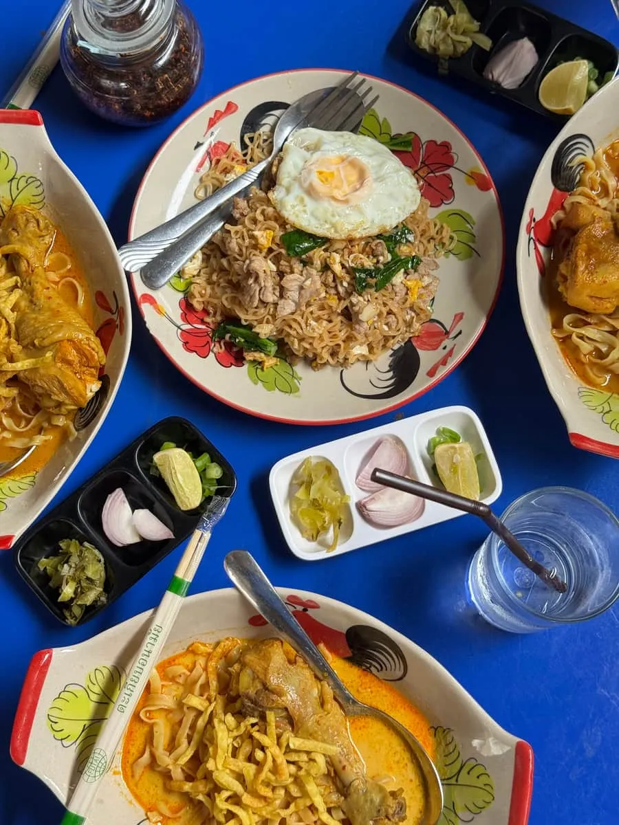 A shared table of khao soi and stir-fried noodles with condiment trays