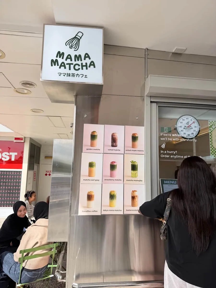 The Mama Matcha shopfront in Hawaiian's Park Centre — the lit whisk-logo sign, the printed drinks board and green chairs with customers seated