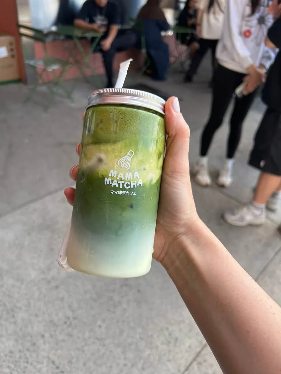A hand holding a Mama Matcha jar outside the Park Centre shopfront, with the queue and green chairs behind